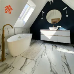 Nowoczesna łazienka na poddaszu z białą wanną wolnostojącą, złotą armaturą, marmurową posadzką i ciemnoniebieską ścianą z lustrem w złotej ramie.