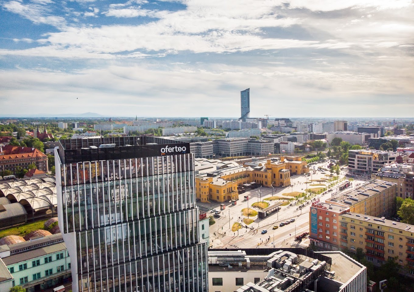 Panorama miasta z nowoczesnym biurowcem z logo Oferteo na dachu, w tle zabytkowy dworzec i drapacz chmur Sky Tower.