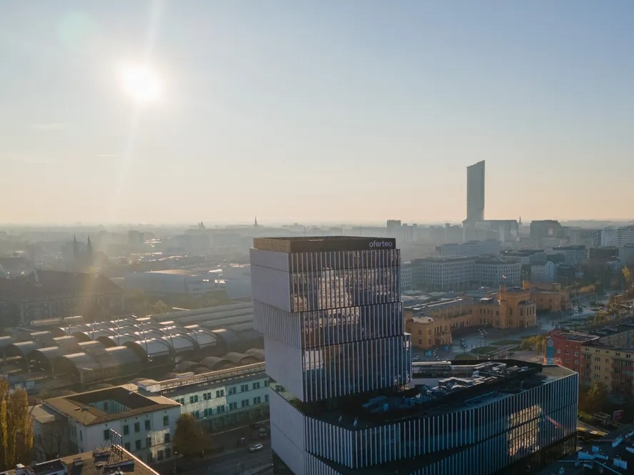 Widok z góry na nowoczesny budynek biurowy z logo Oferteo, w tle panorama miasta w porannym słońcu i mglistej atmosferze.