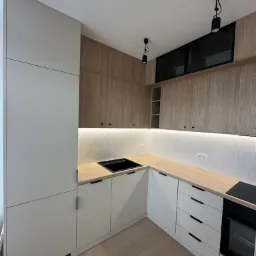 Nowoczesna kuchnia w odcieniach bieli i drewna z czarnymi akcentami, widoczne minimalistyczne szafki górne i dolne, białe płytki ułożone w jodełkę na ścianie oraz drewnopodobna podłoga.