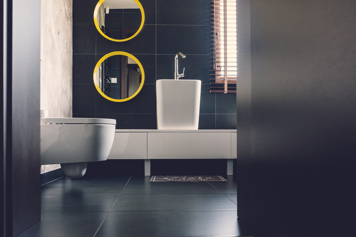 Nowoczesna łazienka z czarnymi płytkami, białym blatem z umywalką i toaletą w stylu minimalistycznym, dwa okrągłe lustra z żółtymi ramami i drewniane żaluzje w oknie.