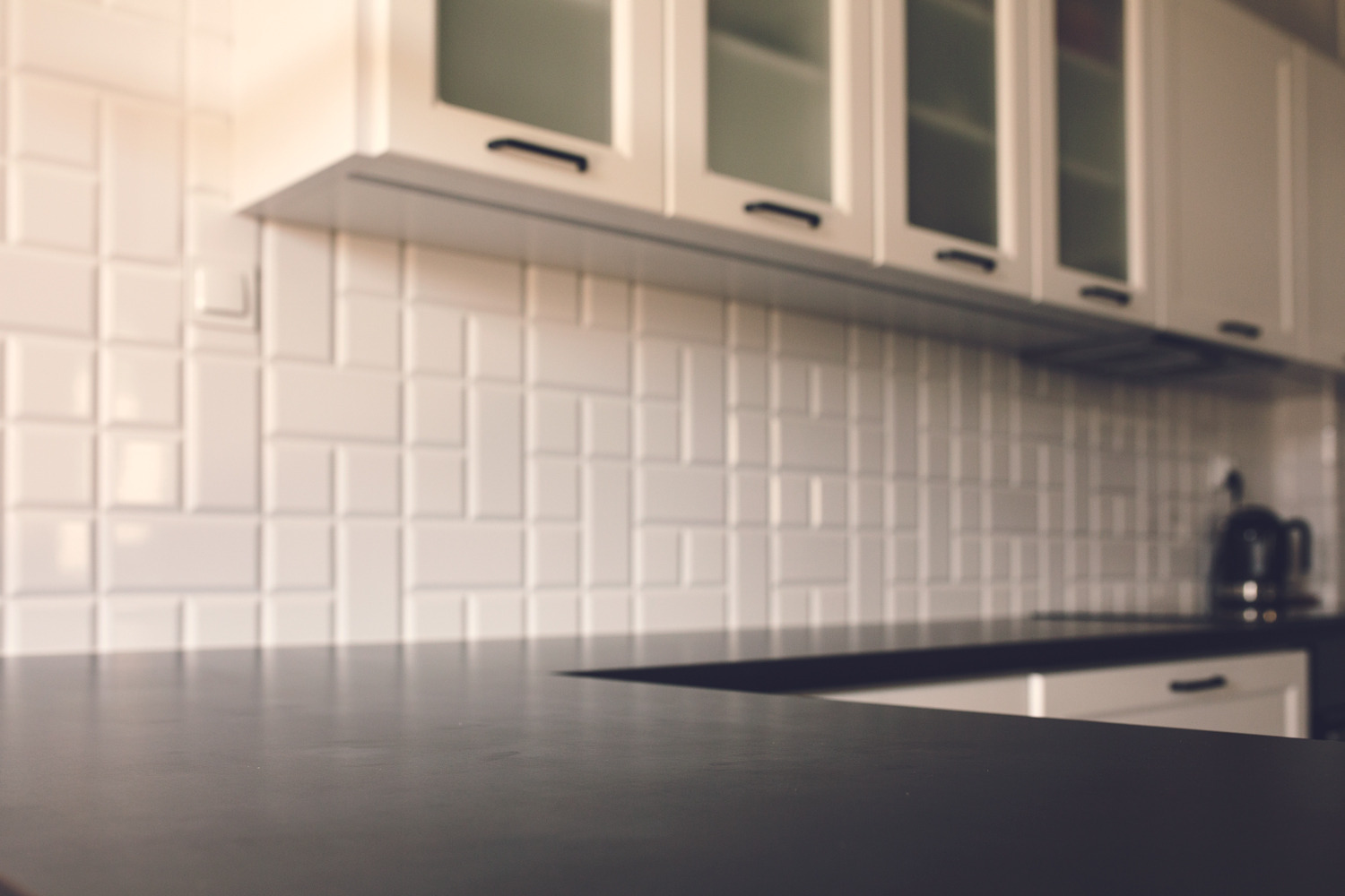 Czarny blat kuchenny z białymi szafkami i płytkami w tle, zlewka z czajnikiem elektrycznym poza głębią ostrości.