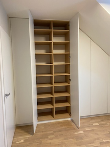 Wysoka, biała szafa wnękowa z otwartymi drzwiami, ukazująca jasne, drewniane półki, dopasowana do skosu sufitu na poddaszu.