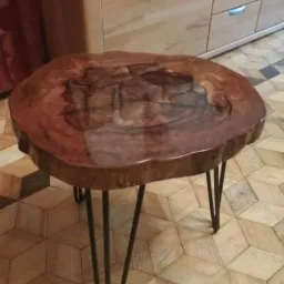 Stolik kawowy z blatem z litego drewna o nieregularnym kształcie, wykończonym żywicą, osadzony na metalowych nogach typu hairpin, na tle parkietu w geometryczny wzór i jasnych mebli.