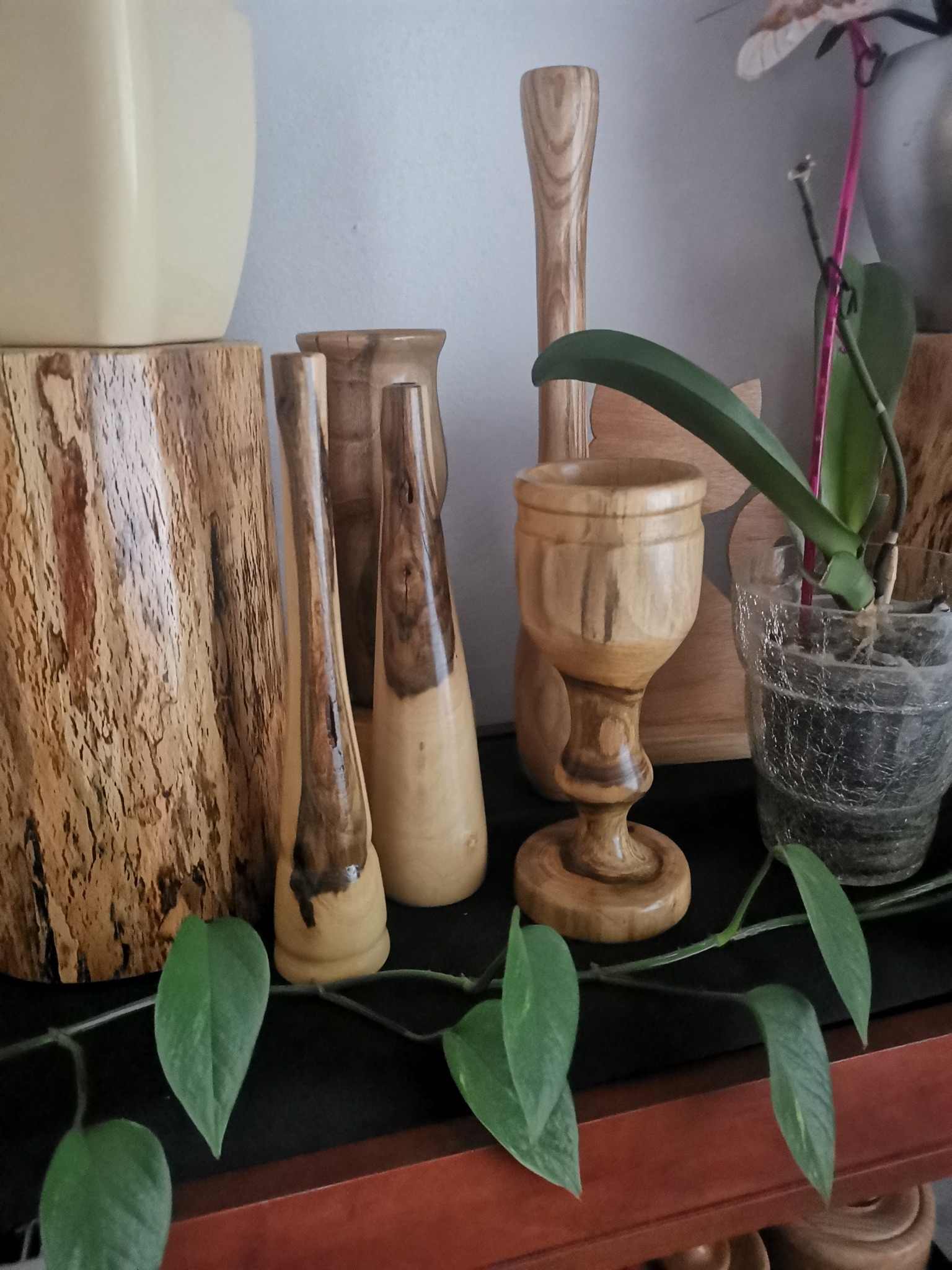 Kolekcja ręcznie toczonych, drewnianych wazonów i kielichów o zróżnicowanych kształtach i odcieniach drewna, ustawiona na półce z roślinami doniczkowymi w tle.