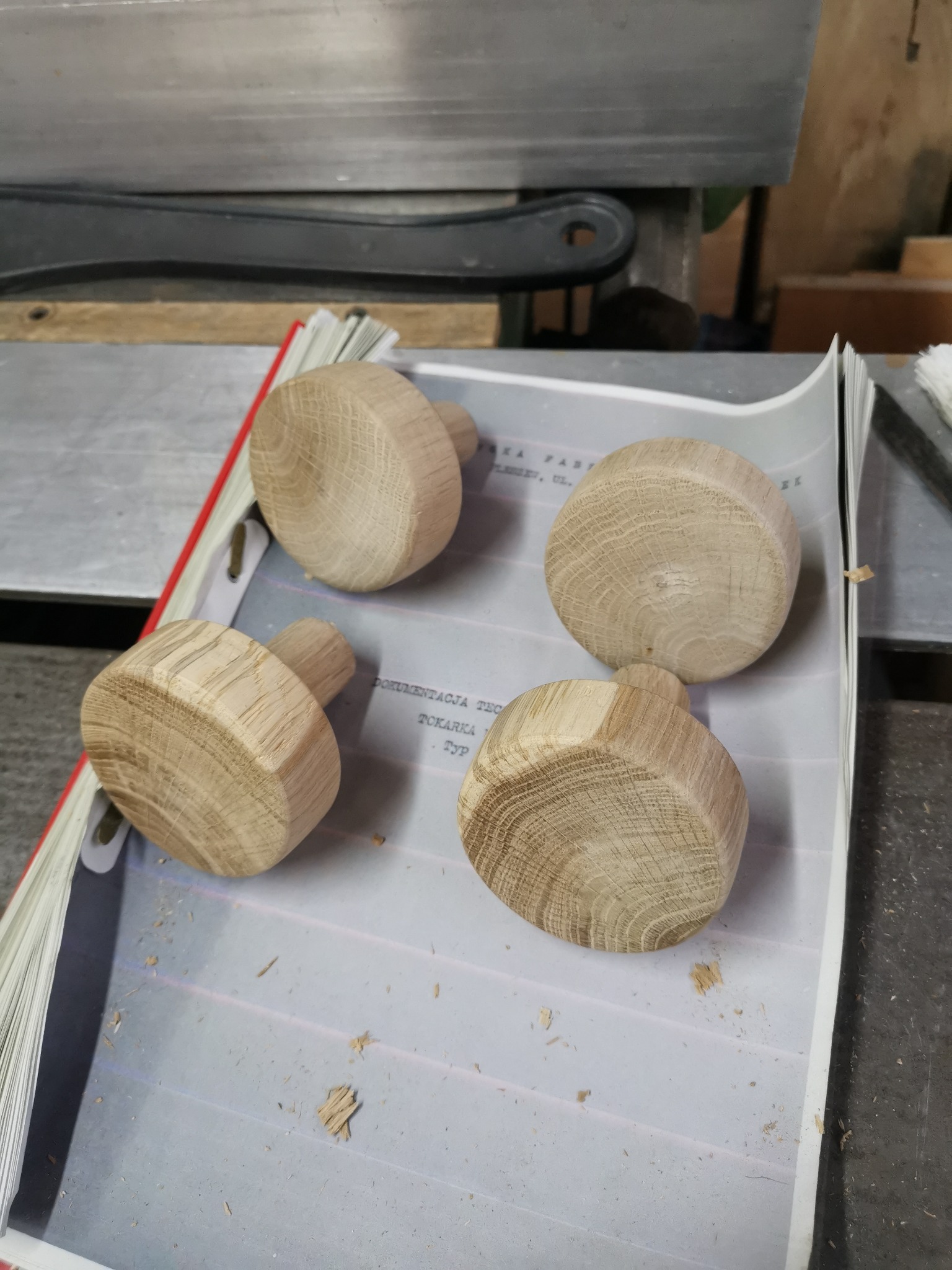 Cztery drewniane gałki z trzonkami, ułożone na otwartej książce z widoczną dokumentacją tokarską, w tle fragment maszyny stolarskiej.