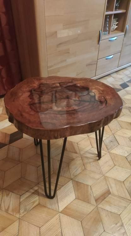 Stolik kawowy z blatem z litego drewna o nieregularnym kształcie, wykończonym żywicą, osadzony na metalowych nogach typu hairpin, na tle parkietu w geometryczny wzór i jasnych mebli.