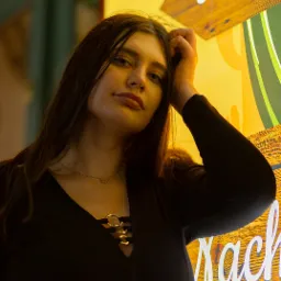 Portret kobiety w czarnej sukience, opierającej się o ścianę z napisem 'Tortillas' i rysunkiem kaktusa w żółto-zielonej kolorystyce, wykonany w nocnej scenerii.