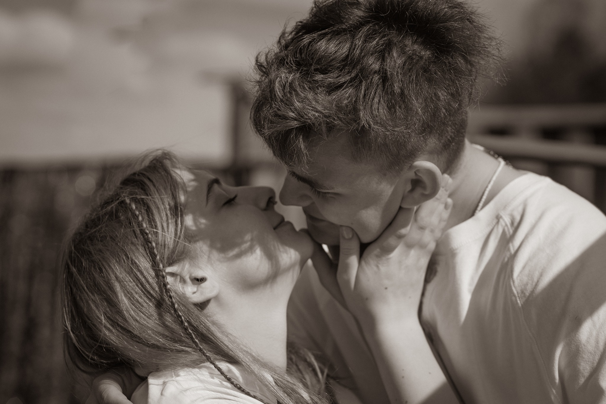 Czarno-białe zbliżenie na parę, delikatnie się całującą; mężczyzna trzyma dłoń na policzku kobiety, ona ma wpięte w warkocz cienkie sznurki.