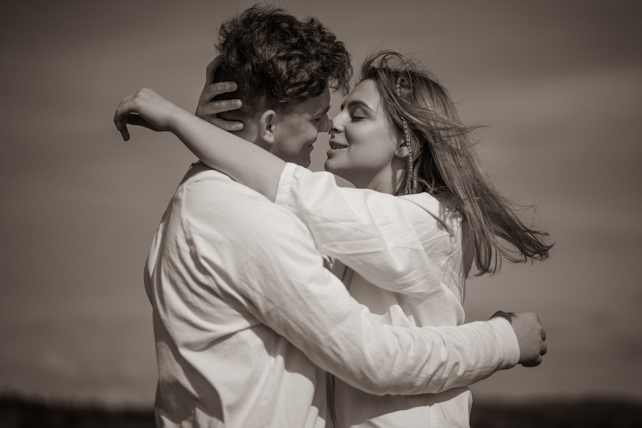 Sepiowe zdjęcie obejmującej się pary w plenerze, ona w koszuli z warkoczykami we włosach, on z kręconymi włosami, z twarzami blisko siebie, na tle nieba i niskiej roślinności.