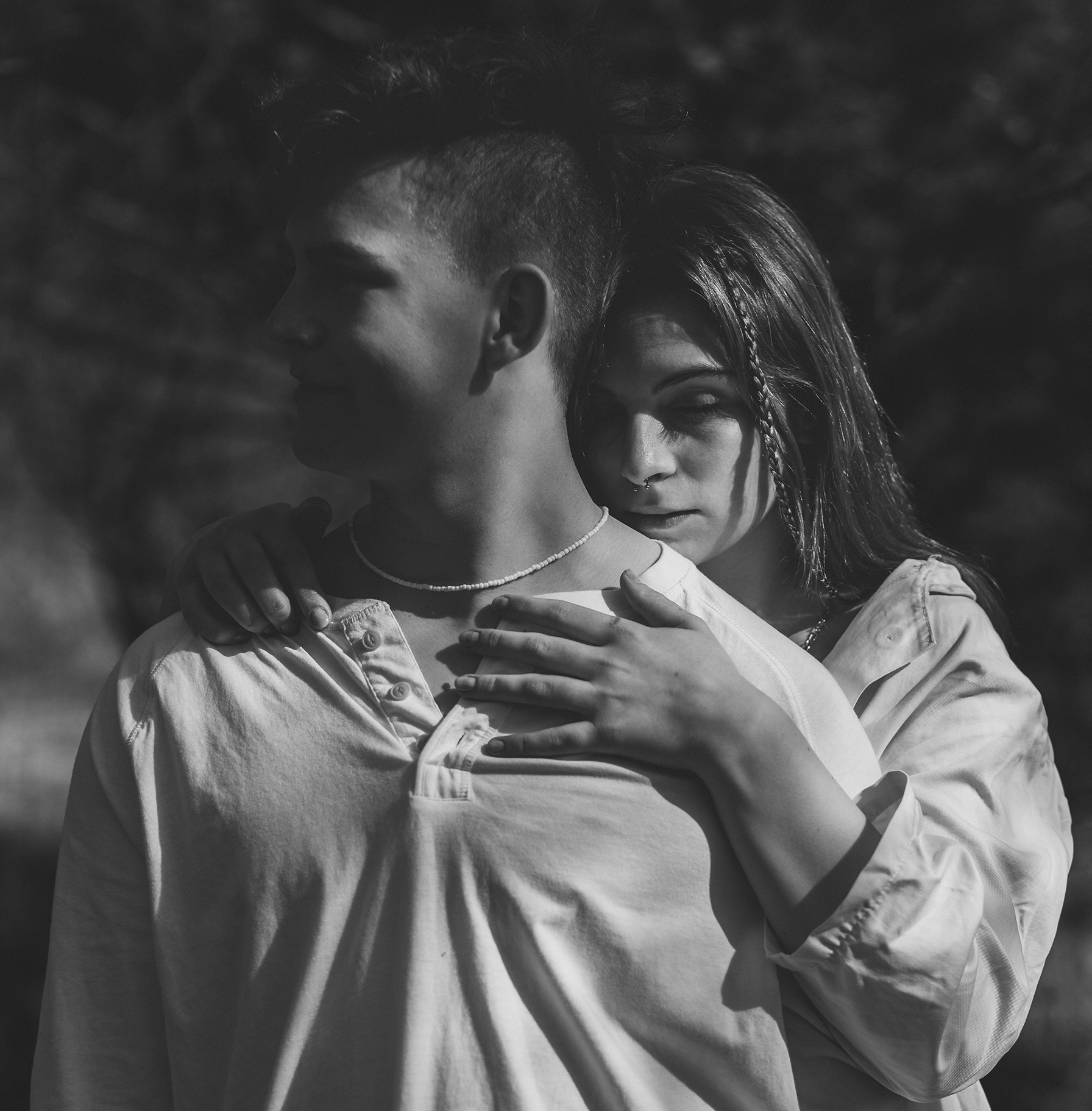 Czarno-białe zbliżenie pary w plenerze: kobieta obejmuje mężczyznę, oboje mają na sobie jasne koszule i naszyjniki z koralików, ona z warkoczykami wplecionymi we włosy, on z krótką fryzurą.