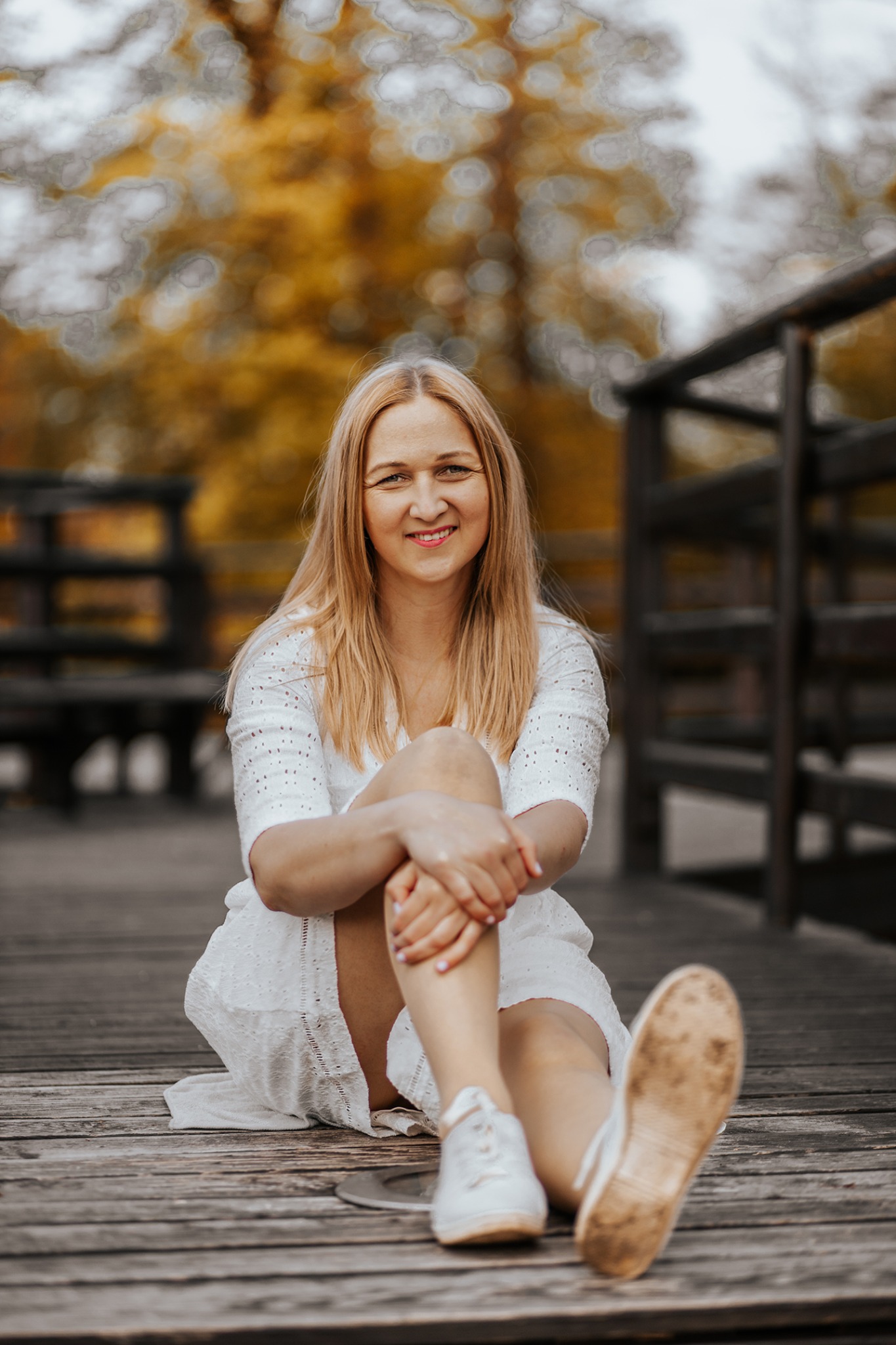 Uśmiechnięta blondynka w białej ażurowej sukience i białych trampkach siedzi na drewnianym pomoście w parku, trzymając kolana i patrząc w obiektyw, jesienne drzewa w tle z efektem bokeh.