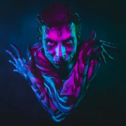 Portret mężczyzny z artystycznym body paintingiem w odcieniach fioletu i błękitu, oświetlonego neonowym światłem, z intensywnym spojrzeniem i rękami skrzyżowanymi na klatce piersiowej.