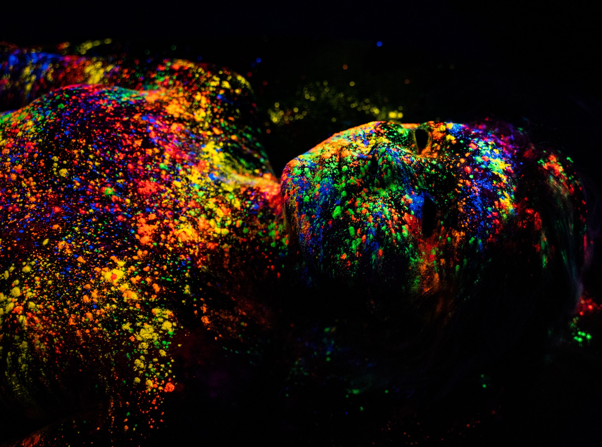 Kobieta leżąca na plecach, pokryta fluorescencyjnymi farbami w odcieniach neonowej zieleni, niebieskiego, pomarańczowego i różowego, oświetlona światłem UV na czarnym tle.