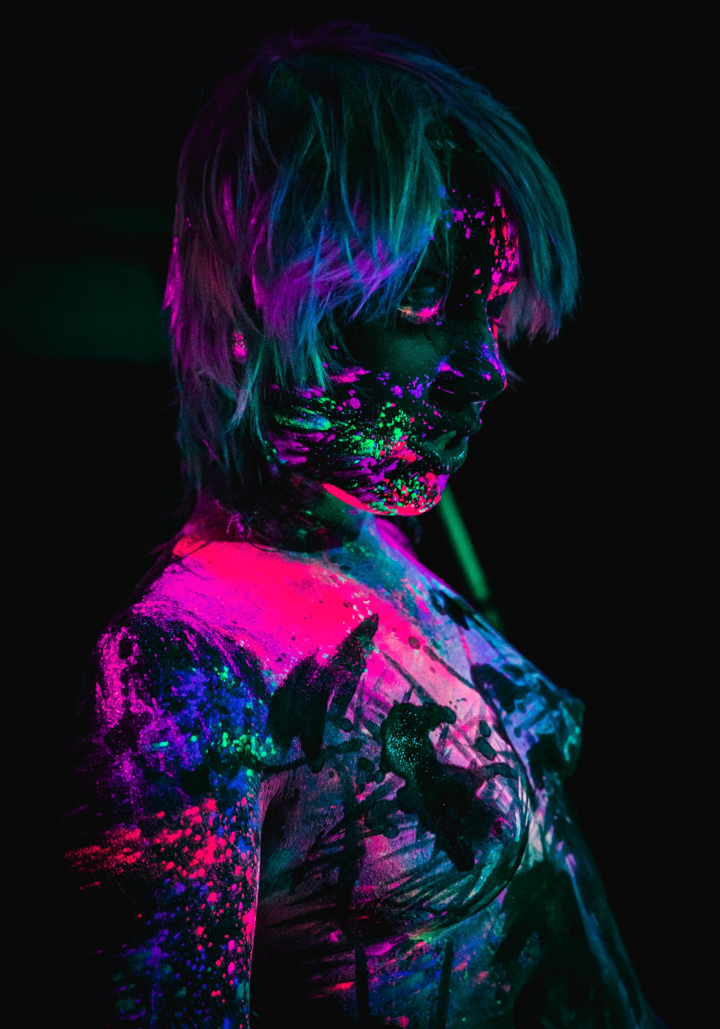 Portret kobiety pokrytej fluorescencyjnymi farbami w odcieniach różu, fioletu, zieleni i błękitu, oświetlonej światłem UV na czarnym tle, z twarzą skierowaną w dół.