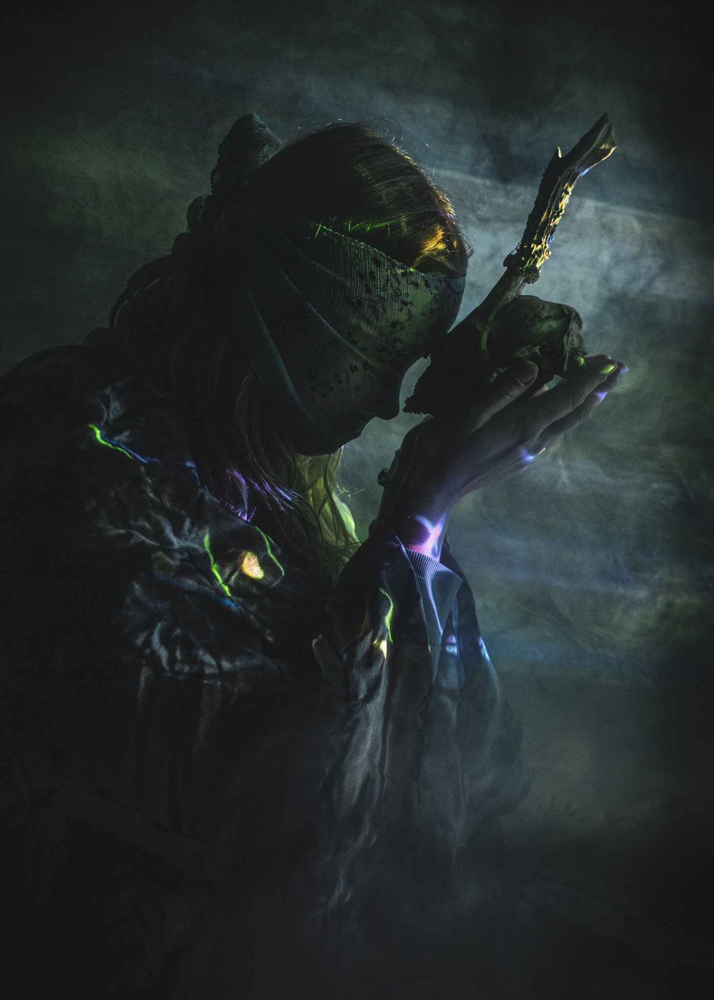 Kobieta w masce i drapowanej szacie trzymająca czaszkę i fragment rogu w dłoniach, oświetlona zielonym i fioletowym światłem w mglistym otoczeniu.