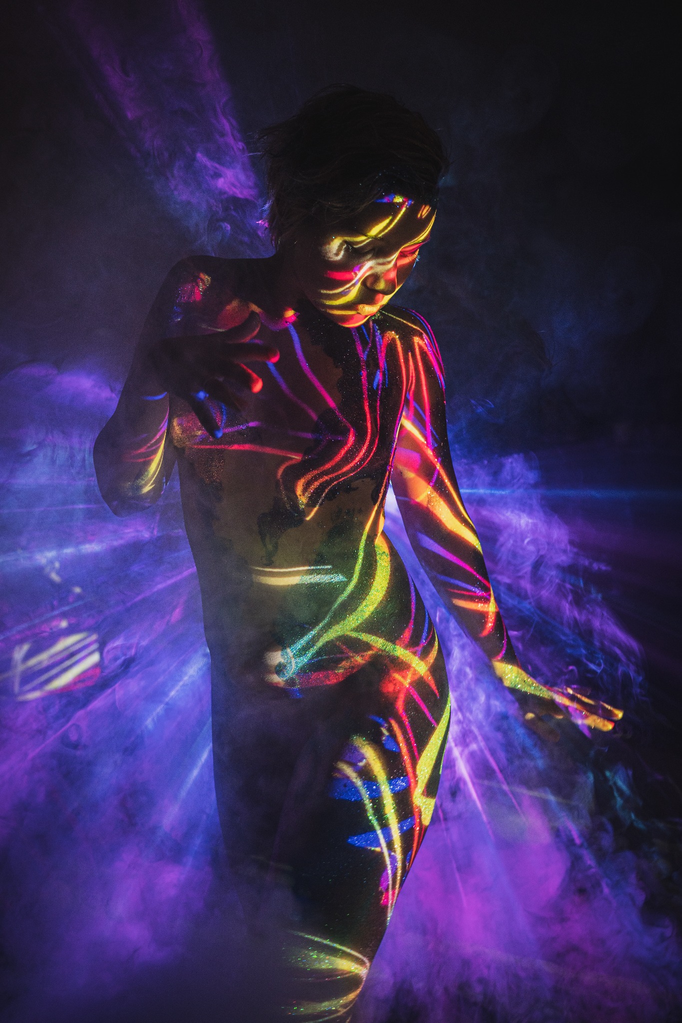 Kobieta w pozującej pozie, ciało pokryte wzorami z kolorowych świateł w otoczeniu fioletowej mgły.