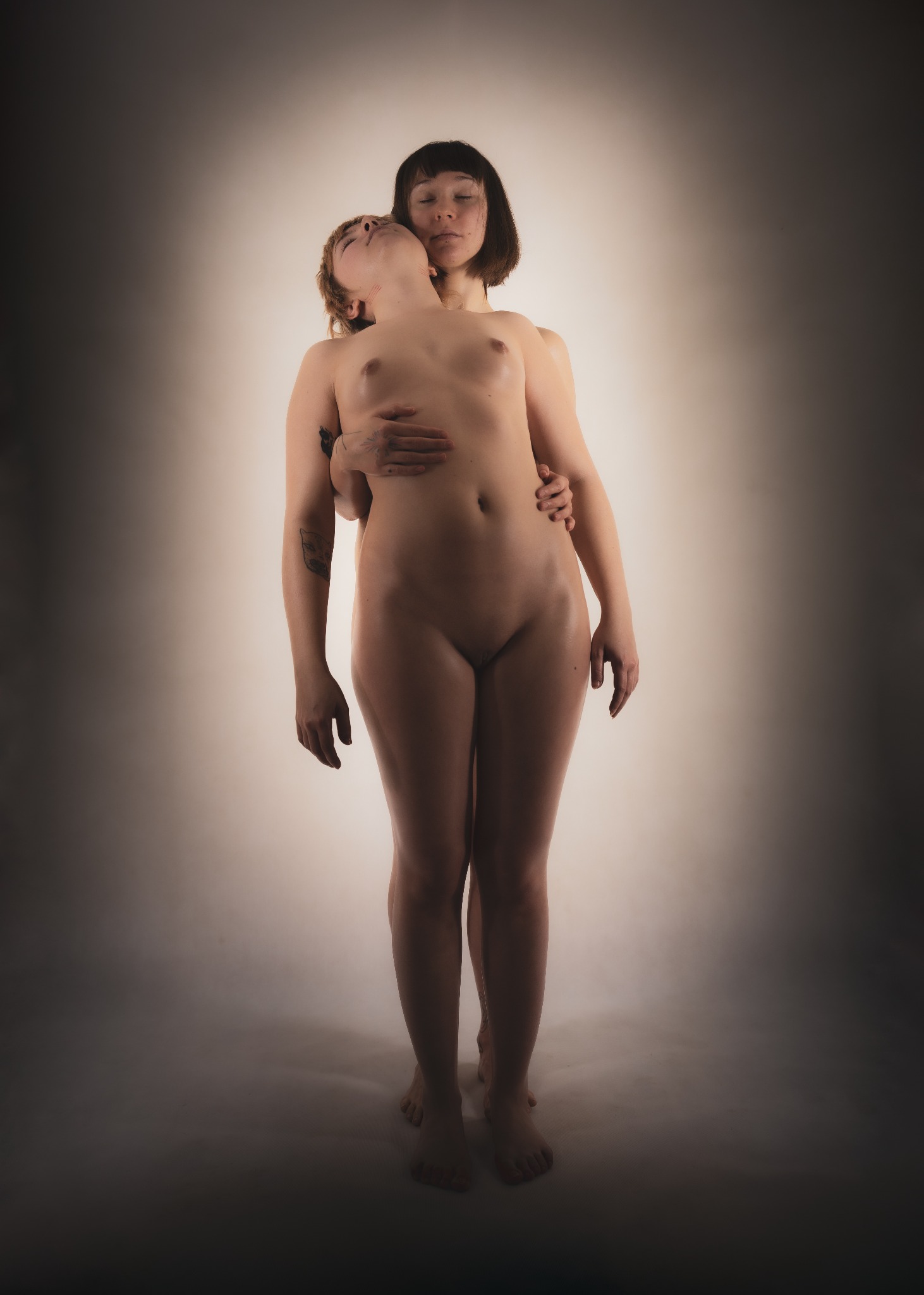 Dwie nagie kobiety w bliskim uścisku na rozmytym, jasnym tle. Jedna kobieta z krótkimi ciemnymi włosami obejmuje drugą, której głowa jest odchylona do tyłu, a na ramieniu widać tatuaż.
