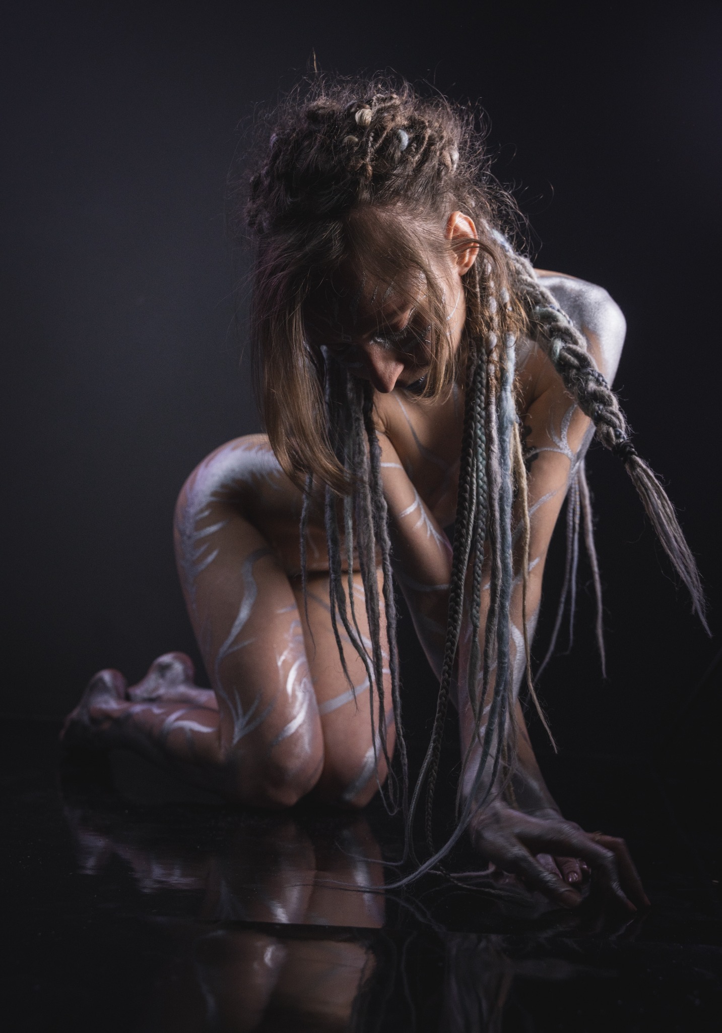 Kobieta w pozycji klęczącej, z dredami i srebrnymi wzorami na ciele, w ciemnym studio, delikatne odbicie na podłodze, artystyczne ujęcie.