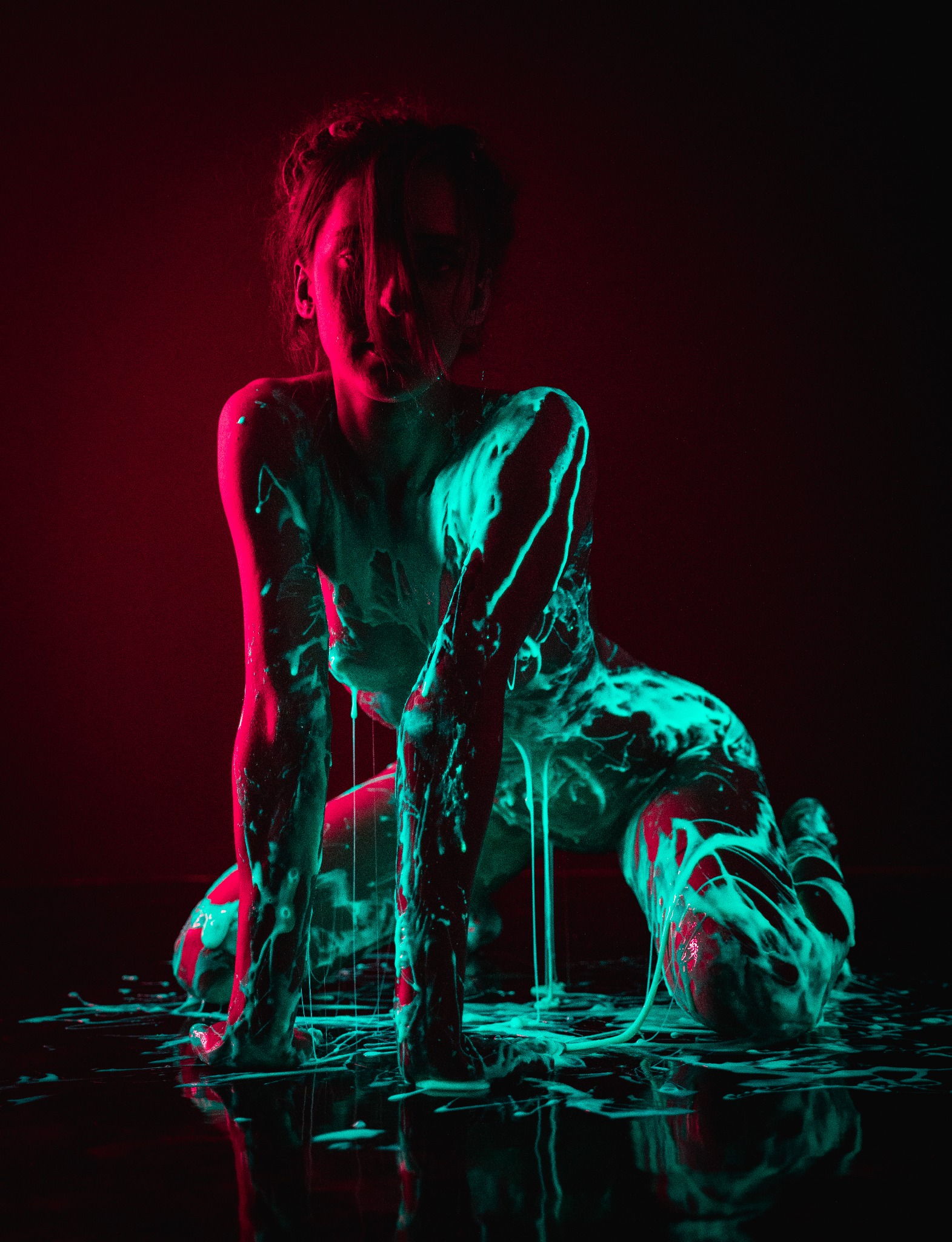 Kobieta na czworakach, pokryta białą substancją, oświetlona czerwonym i turkusowym światłem, z widocznymi smugami substancji na podłodze.