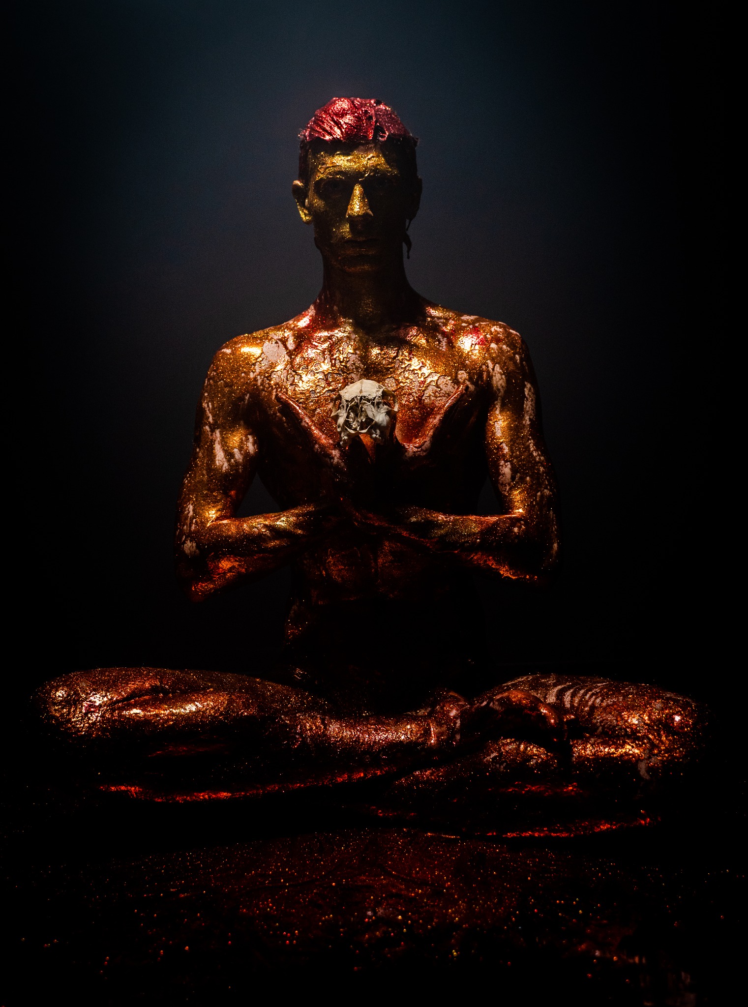 Mężczyzna w pozycji lotosu, ciało pokryte złotym brokatem, trzymający małą czaszkę w dłoniach, czerwone włosy, ciemne tło.