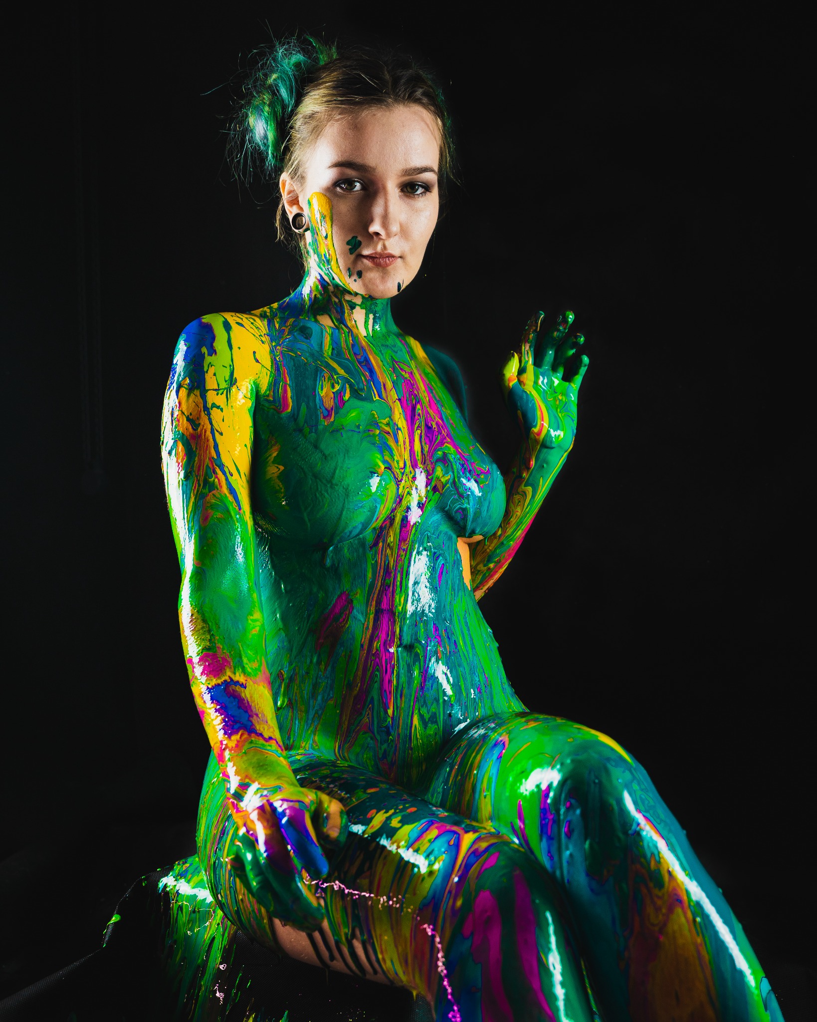 Portret kobiety w całości pokrytej wielobarwną farbą, z zielonymi pasemkami we włosach, pozującej na czarnym tle, z uniesioną dłonią.