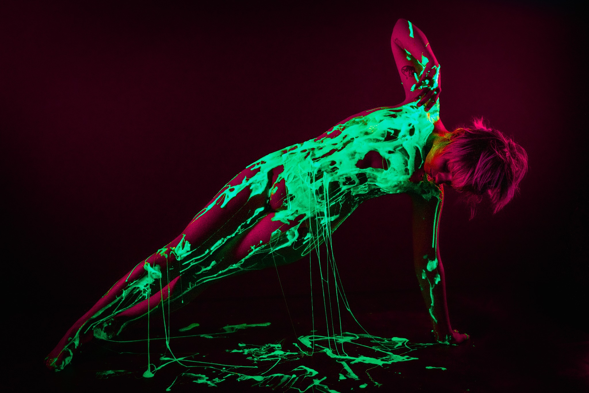Kobieta w pozycji bocznej deski, pokryta świecącą w ciemności farbą w odcieniach zieleni na tle ciemnego, purpurowego tła, tworząca abstrakcyjny i artystyczny efekt.