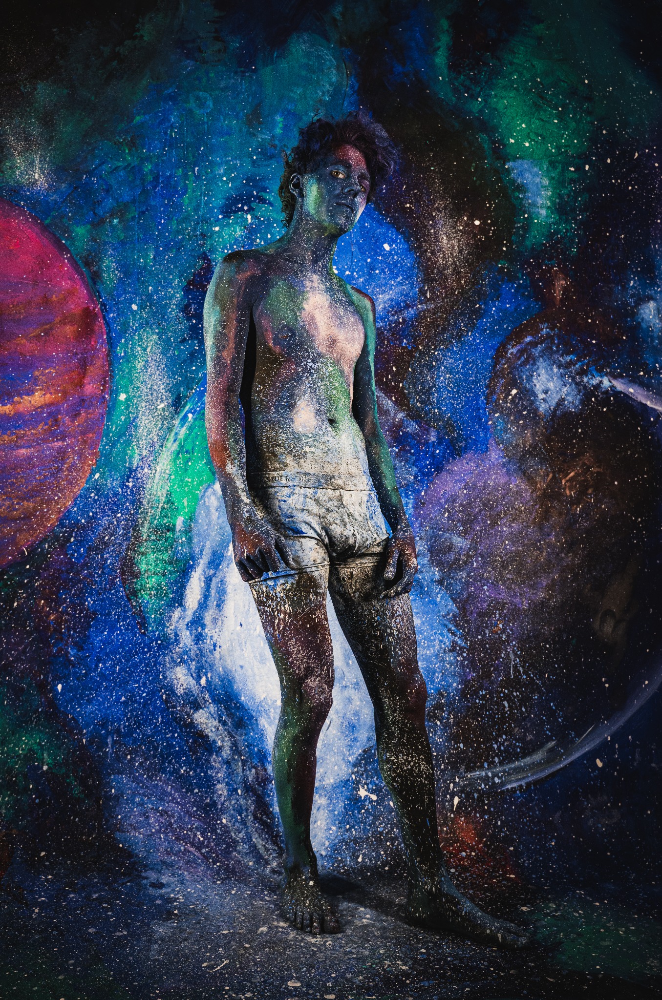Mężczyzna pokryty farbą w kolorach kosmosu, pozujący na tle galaktycznej scenografii, z delikatnym oświetleniem podkreślającym fakturę farby na jego ciele.