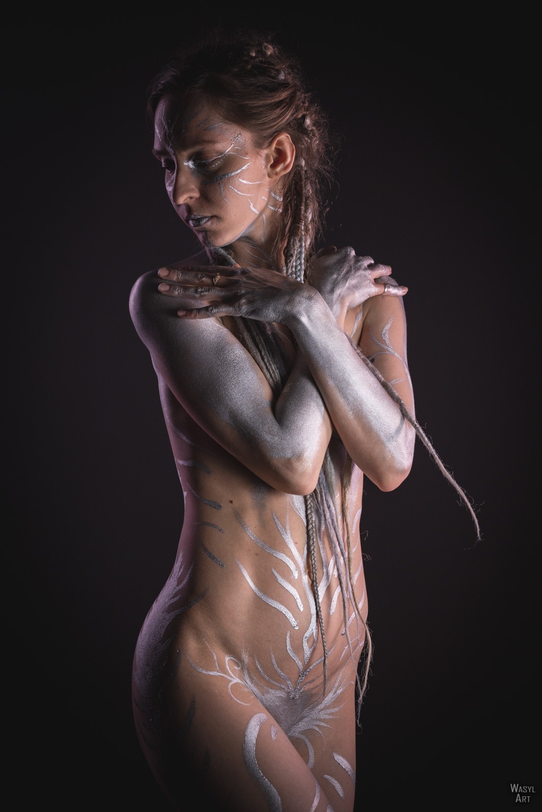 Kobieta z artystycznym, srebrnym body paintingiem w kształcie roślinnych motywów, włosy zaplecione w warkocze, pozuje na ciemnym tle