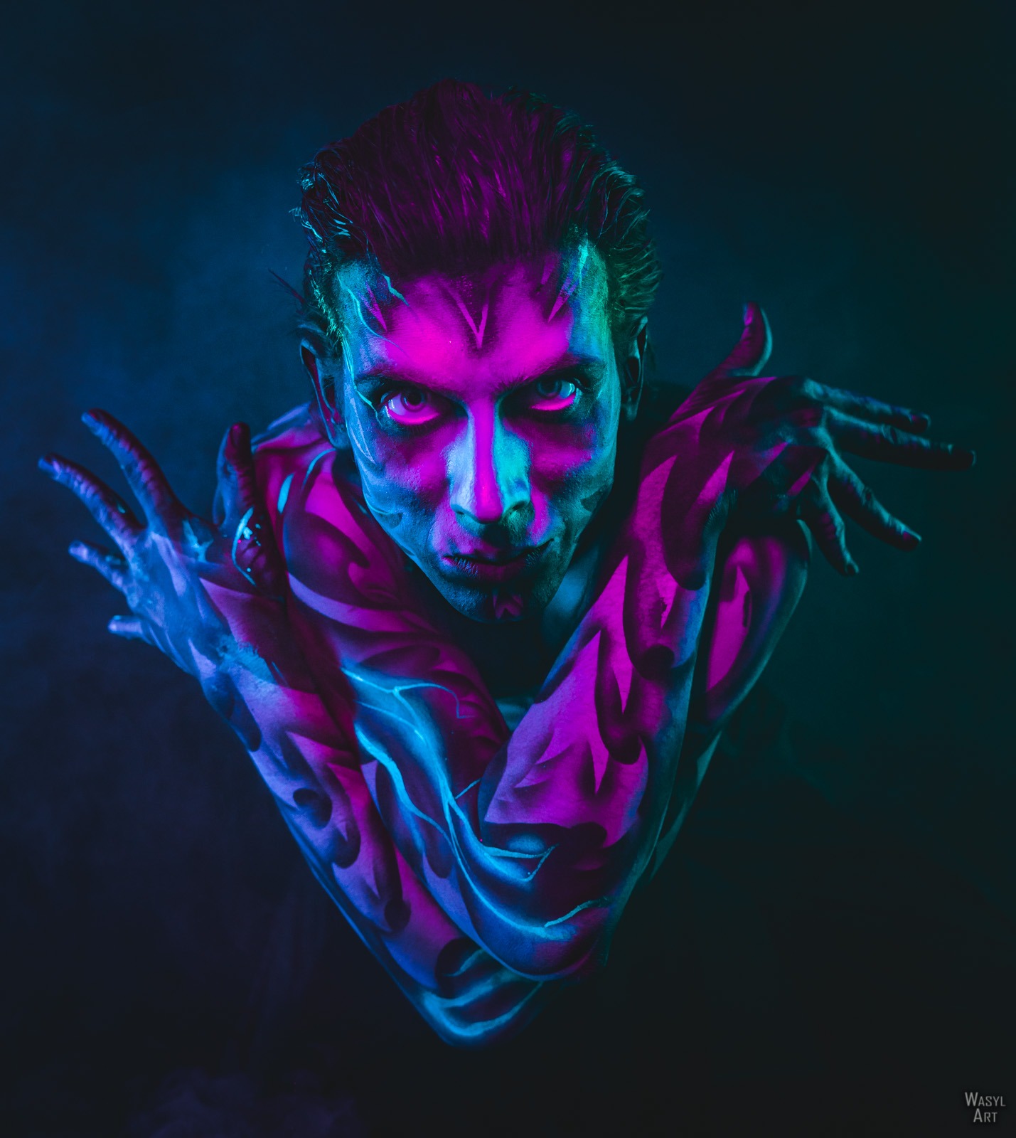 Portret mężczyzny z artystycznym body paintingiem w odcieniach fioletu i błękitu, oświetlonego neonowym światłem, z intensywnym spojrzeniem i rękami skrzyżowanymi na klatce piersiowej.