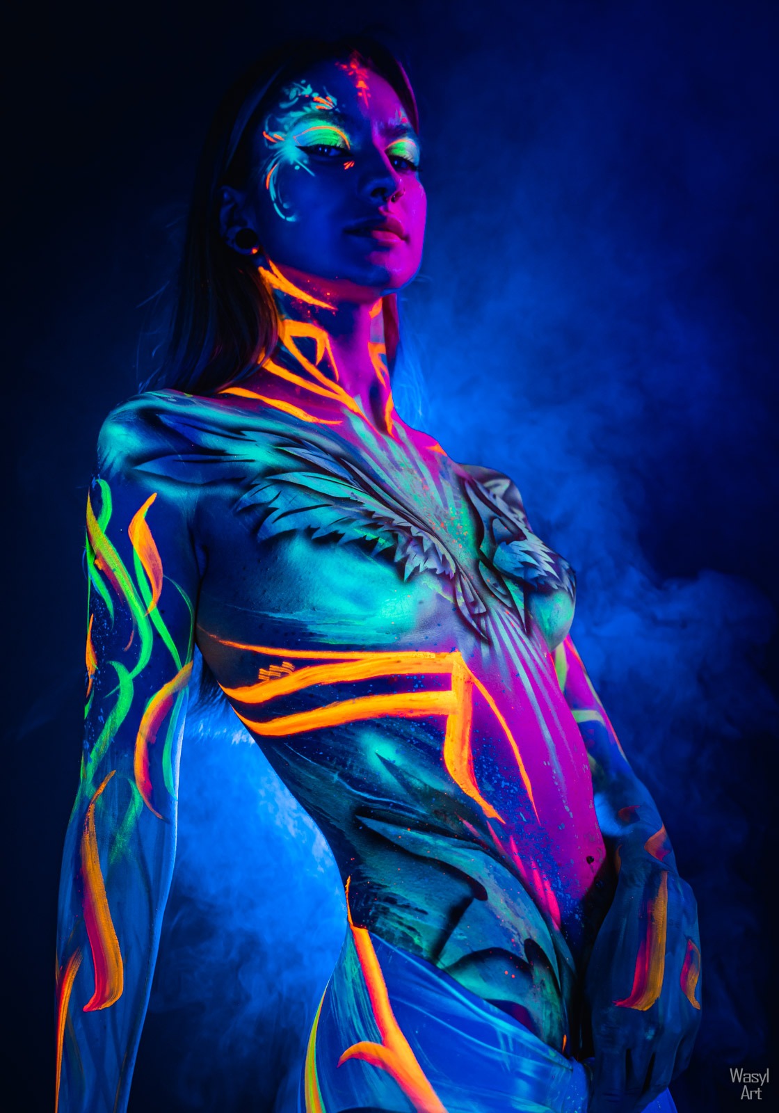 Portret kobiety z artystycznym body paintingiem fluorescencyjnym w odcieniach neonowej zieleni, pomarańczy i różu na tle ciemnego dymu.