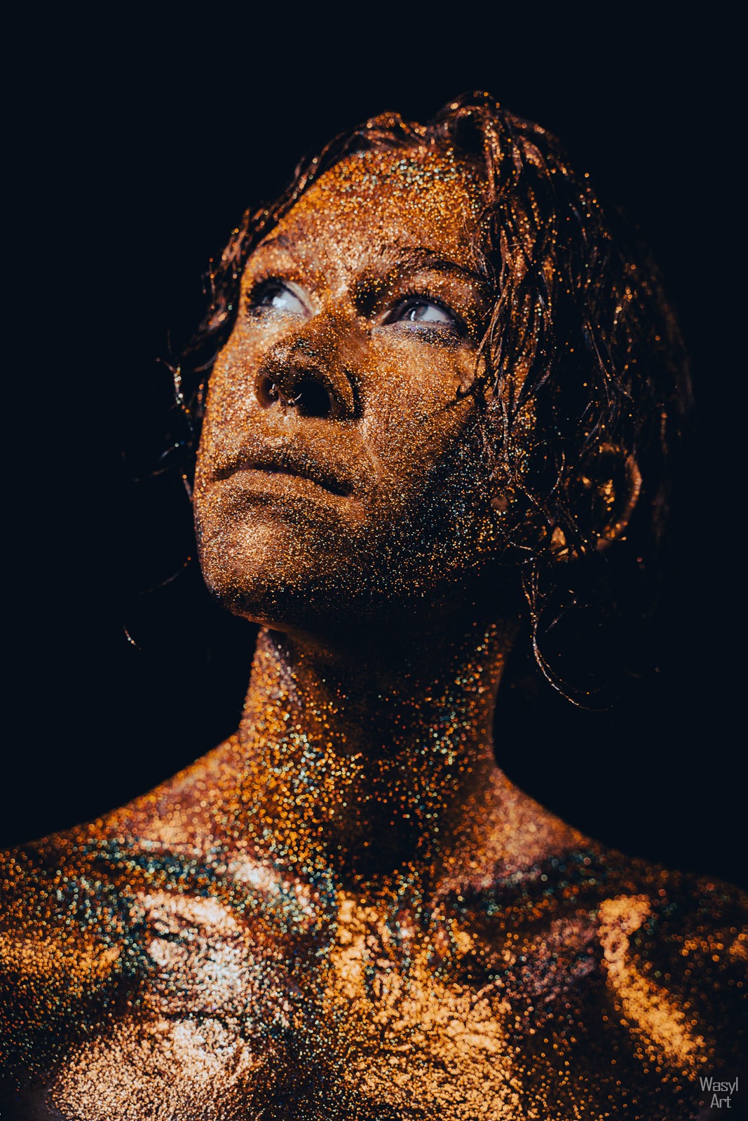 Portret kobiety z ciałem pokrytym złotym brokatem, spoglądającej w górę na ciemnym tle.
