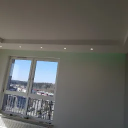 Jasne wnętrze po remoncie: gładkie ściany, biały sufit z oświetleniem punktowym, okno z widokiem na miasto.