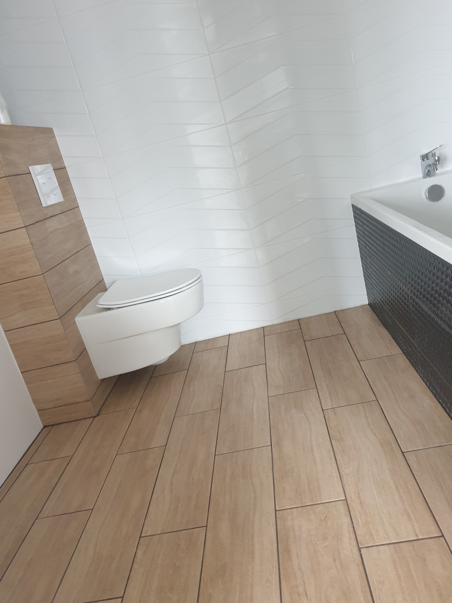 Nowoczesna łazienka z podwieszaną toaletą, białymi płytkami ściennymi z geometrycznym wzorem i brązowymi płytkami podłogowymi imitującymi drewno, obudowa wanny z dekoracyjną fakturą.