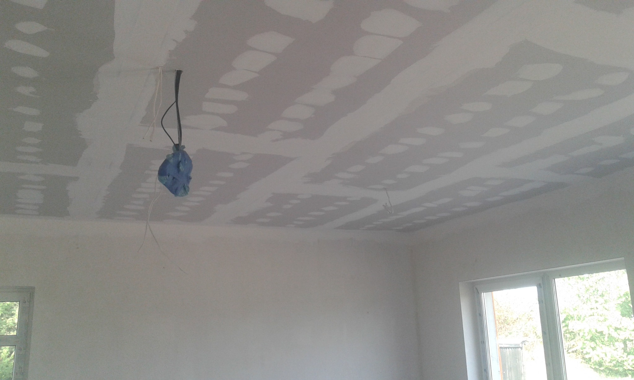 Sufit po nałożeniu pierwszej warstwy szpachli, widoczne łączenia płyt gipsowo-kartonowych, zabezpieczone kable zwisają z otworu, okno w tle.