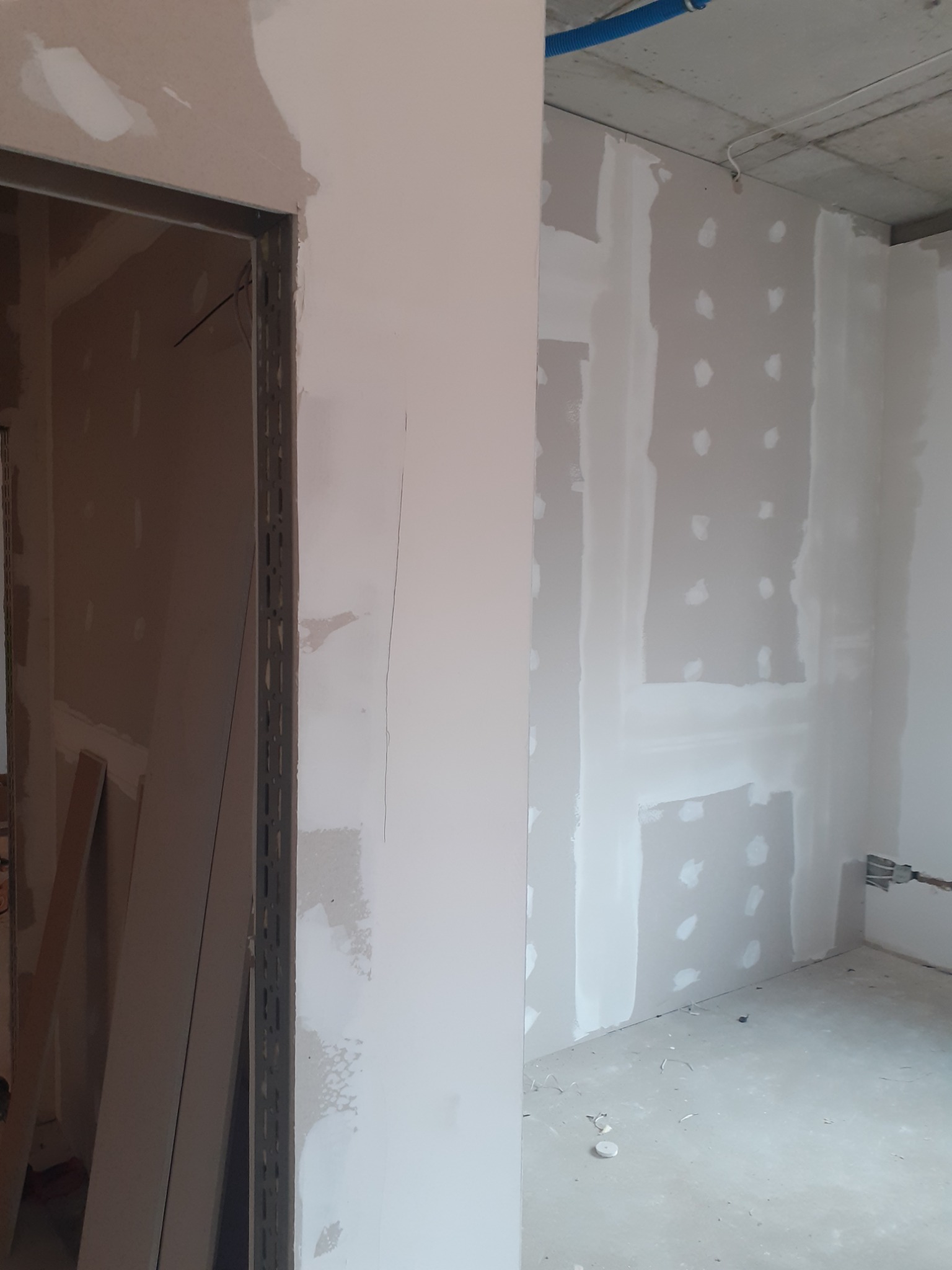 Ściana w trakcie prac przygotowawczych do wykończenia, widoczne punktowe nakładanie masy szpachlowej na płyty gipsowo-kartonowe, metalowa ościeżnica drzwiowa, surowy betonowy sufit z instalacją.