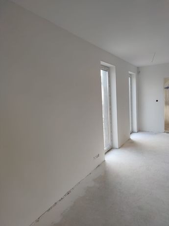 Wnętrze w stanie deweloperskim z gładkimi, białymi ścianami i surową betonową podłogą; widoczne dwa okna z białymi ramami i wystającymi parapetami.