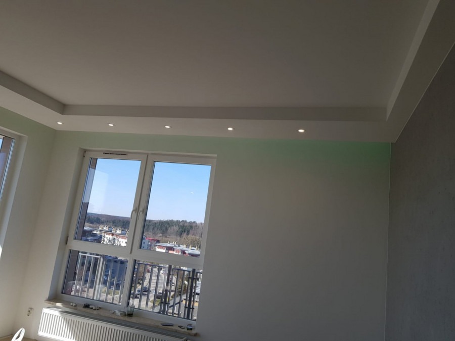 Jasne wnętrze po remoncie: gładkie ściany, biały sufit z oświetleniem punktowym, okno z widokiem na miasto.