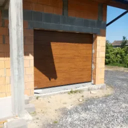 Brązowa brama segmentowa z poziomym wzorem drewna w surowym otworze garażowym z cegły, widoczny betonowy słup i gruz na podjeździe.