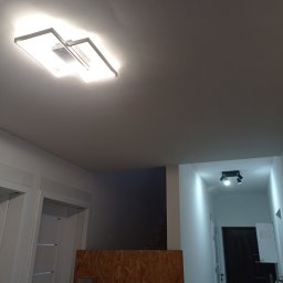 Usługi Budowlane Krzysztof Sandrzyk - Nowoczesna lampa sufitowa LED w kształcie kwadratu, oświetlająca wnętrze w trakcie prac wykończeniowych; widoczne zabezpieczenia z płyty OSB i fragment korytarza z dodatkowym oświetleniem punktowym.
