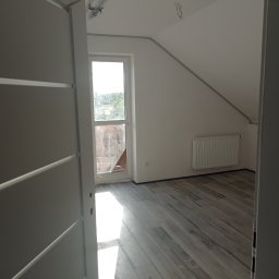 Usługi Budowlane Krzysztof Sandrzyk - Poddasze po remoncie z panelami podłogowymi imitującymi drewno, widok przez otwarte drzwi z białymi, poziomymi panelami dekoracyjnymi, grzejnik na ścianie, okno dachowe z widokiem na zewnątrz.