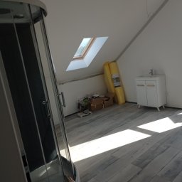 Usługi Budowlane Krzysztof Sandrzyk - Poddasze w trakcie prac wykończeniowych: częściowo ułożona podłoga z paneli, kabina prysznicowa z widocznymi narzędziami, biała szafka z umywalką i zwinięte panele pod ścianą.