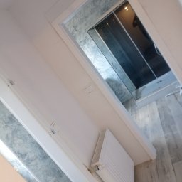 Usługi Budowlane Krzysztof Sandrzyk - Wykończona łazienka z kabiną prysznicową, grzejnikiem na ścianie i podłogą imitującą drewno, widziana z perspektywy wnętrza mieszkania.