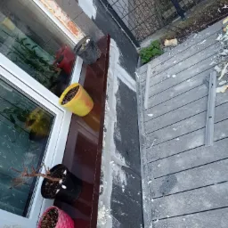 Widok z góry na okno z doniczkami podczas prac wykończeniowych elewacji, z widoczną pianką montażową i deskami tarasowymi na zewnątrz.