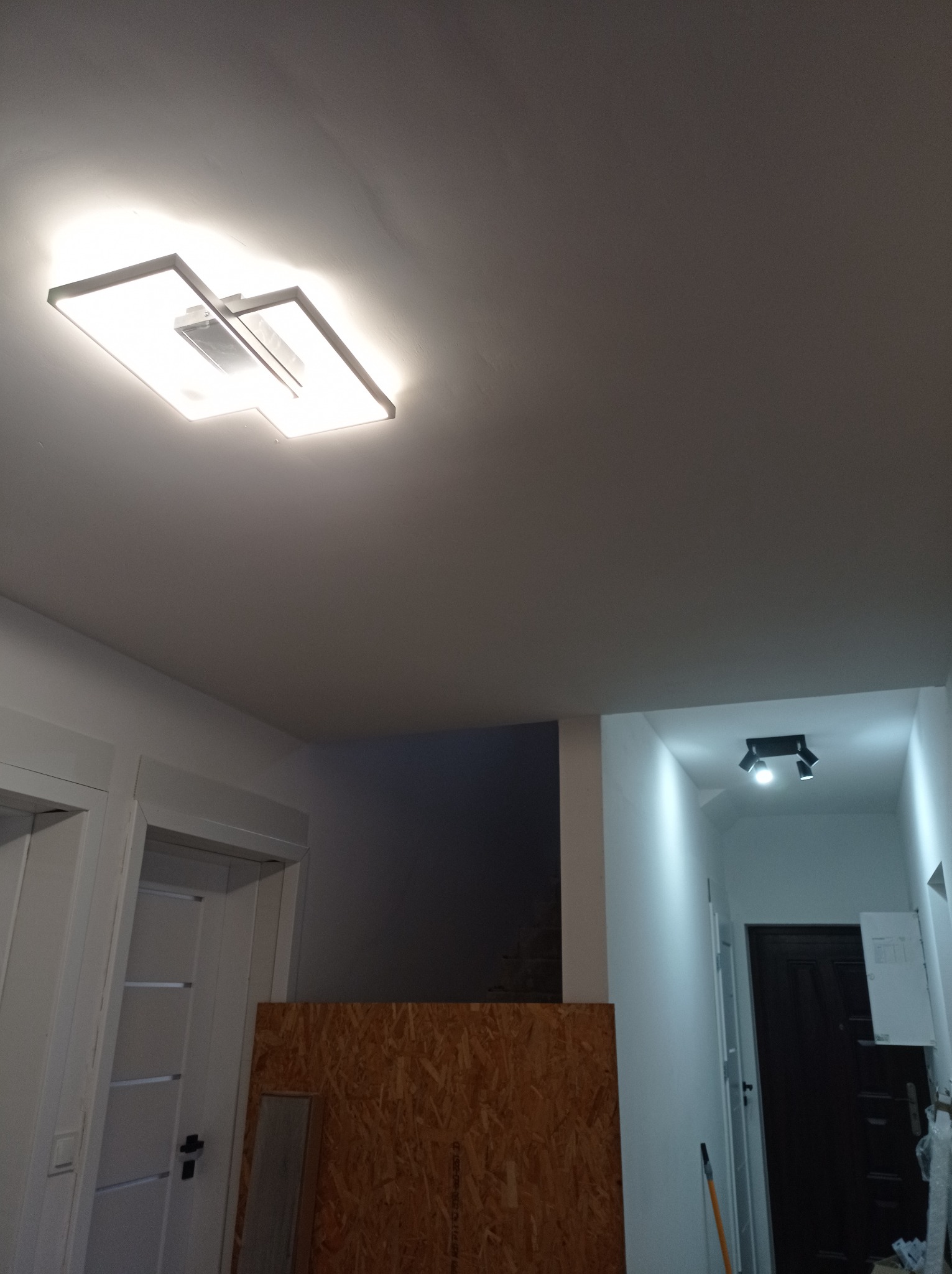 Nowoczesna lampa sufitowa LED w kształcie kwadratu, oświetlająca wnętrze w trakcie prac wykończeniowych; widoczne zabezpieczenia z płyty OSB i fragment korytarza z dodatkowym oświetleniem punktowym.