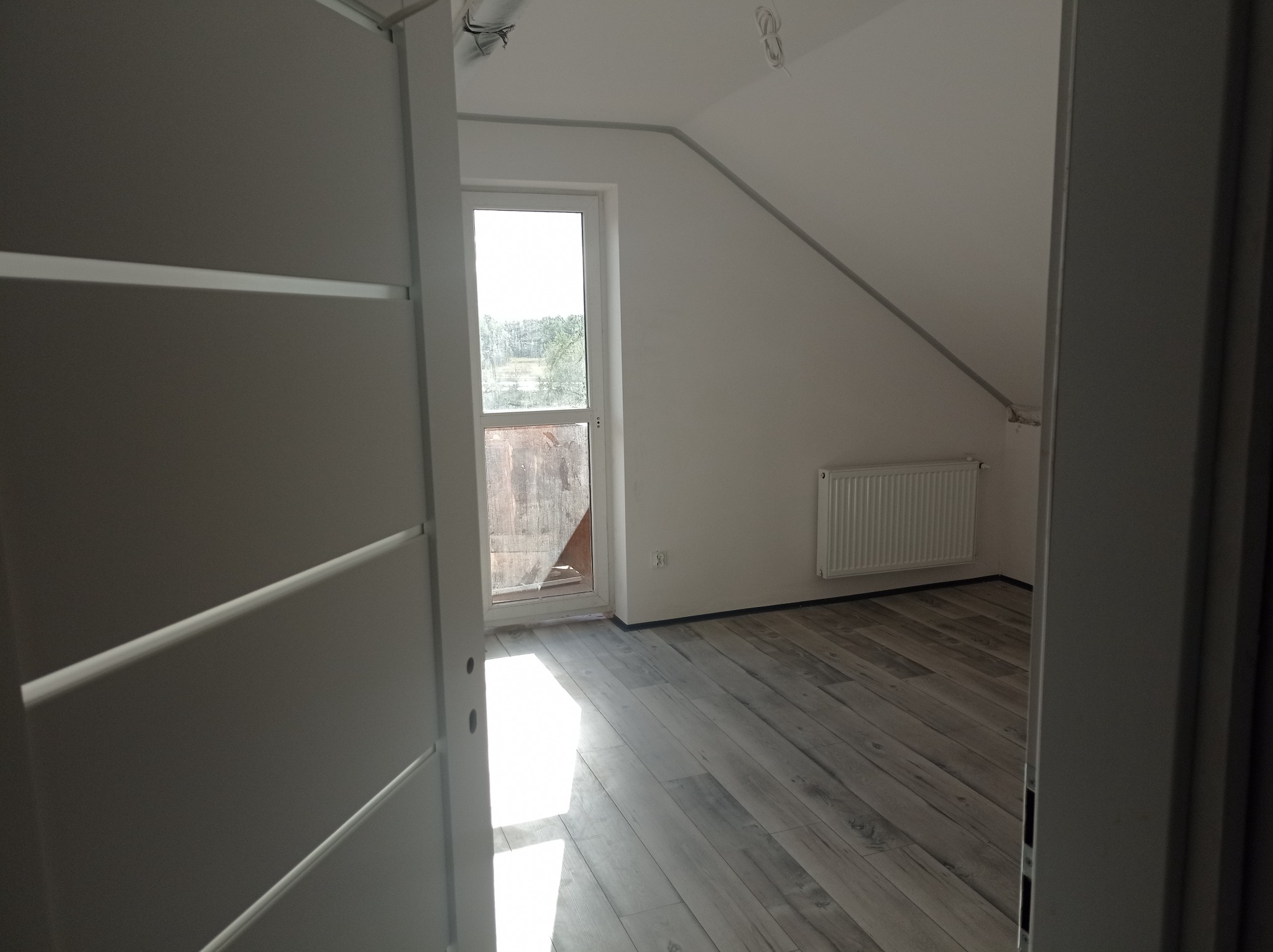 Poddasze po remoncie z panelami podłogowymi imitującymi drewno, widok przez otwarte drzwi z białymi, poziomymi panelami dekoracyjnymi, grzejnik na ścianie, okno dachowe z widokiem na zewnątrz.