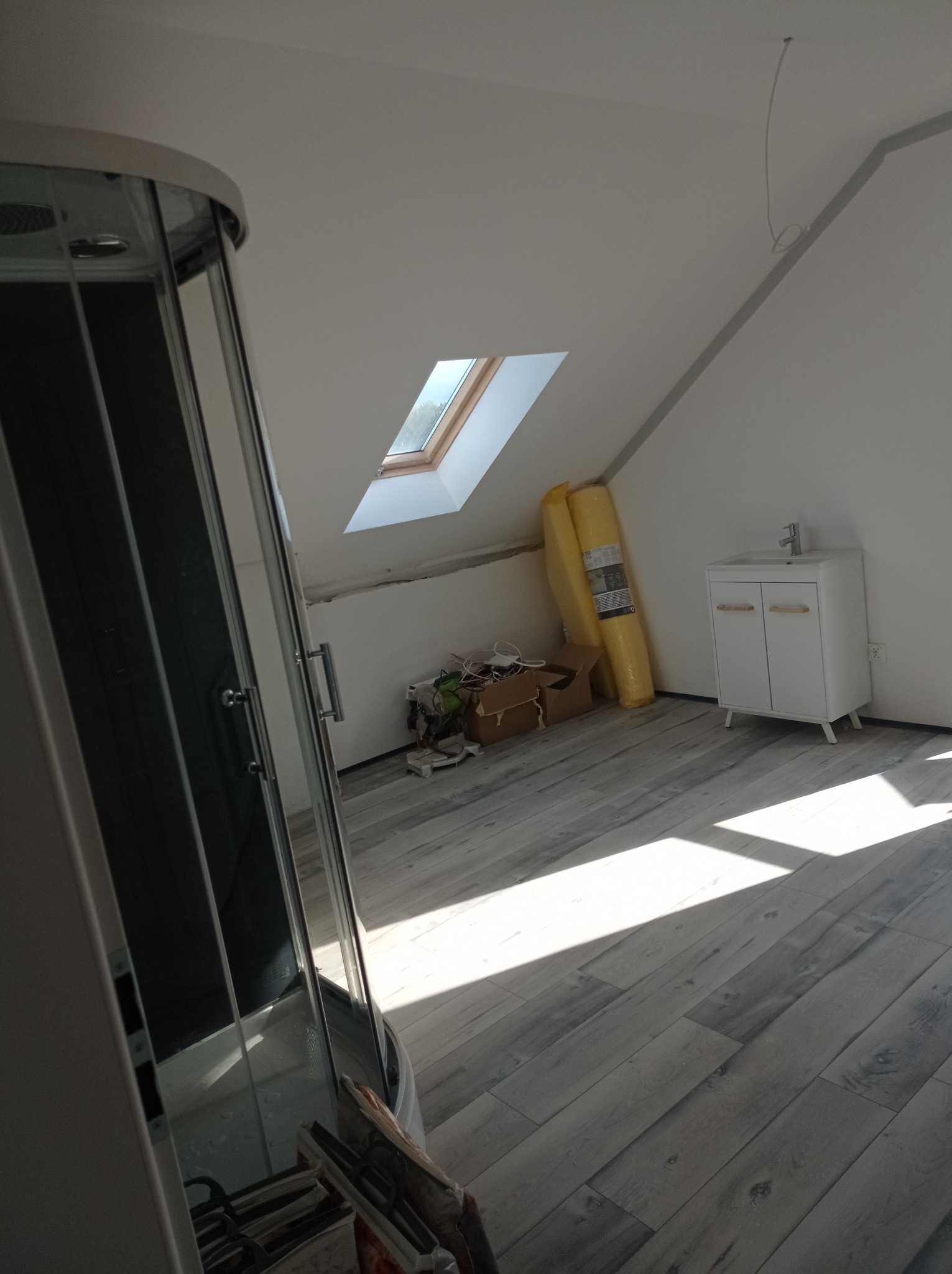 Poddasze w trakcie prac wykończeniowych: częściowo ułożona podłoga z paneli, kabina prysznicowa z widocznymi narzędziami, biała szafka z umywalką i zwinięte panele pod ścianą.