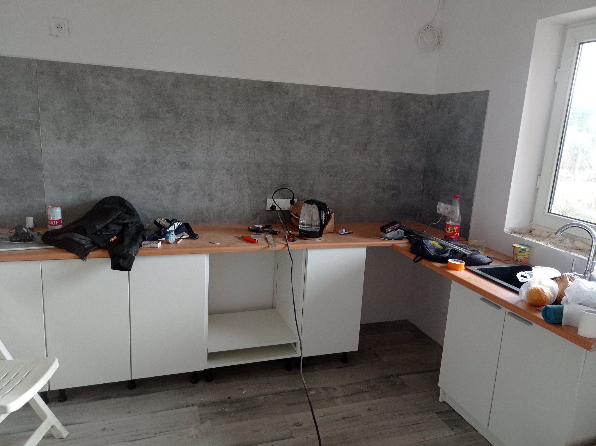 Wnętrze kuchni w trakcie montażu mebli: białe szafki z drewnianym blatem, szara ściana, widoczne narzędzia i akcesoria, podłoga wyłożona panelami imitującymi drewno.