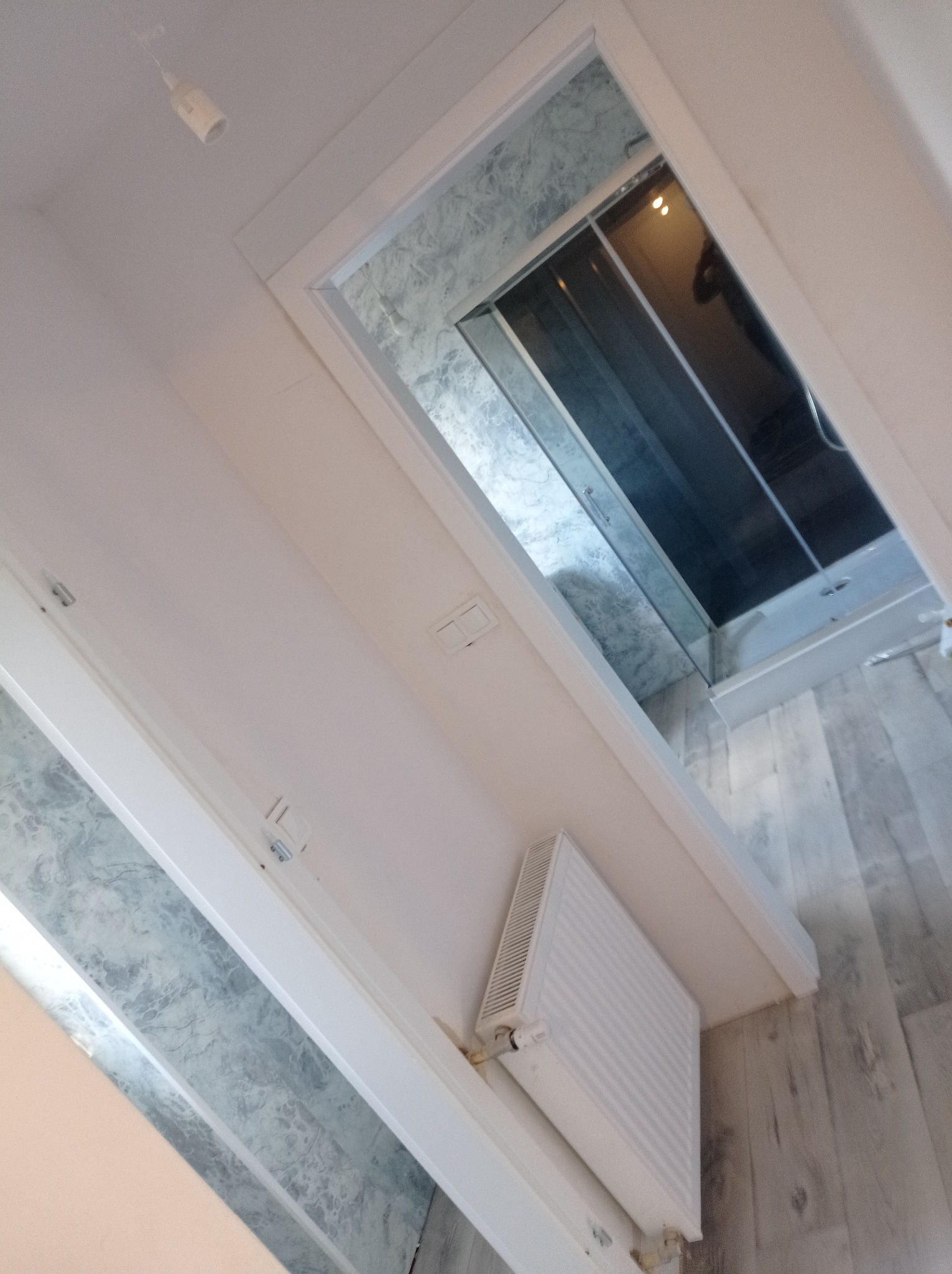 Wykończona łazienka z kabiną prysznicową, grzejnikiem na ścianie i podłogą imitującą drewno, widziana z perspektywy wnętrza mieszkania.