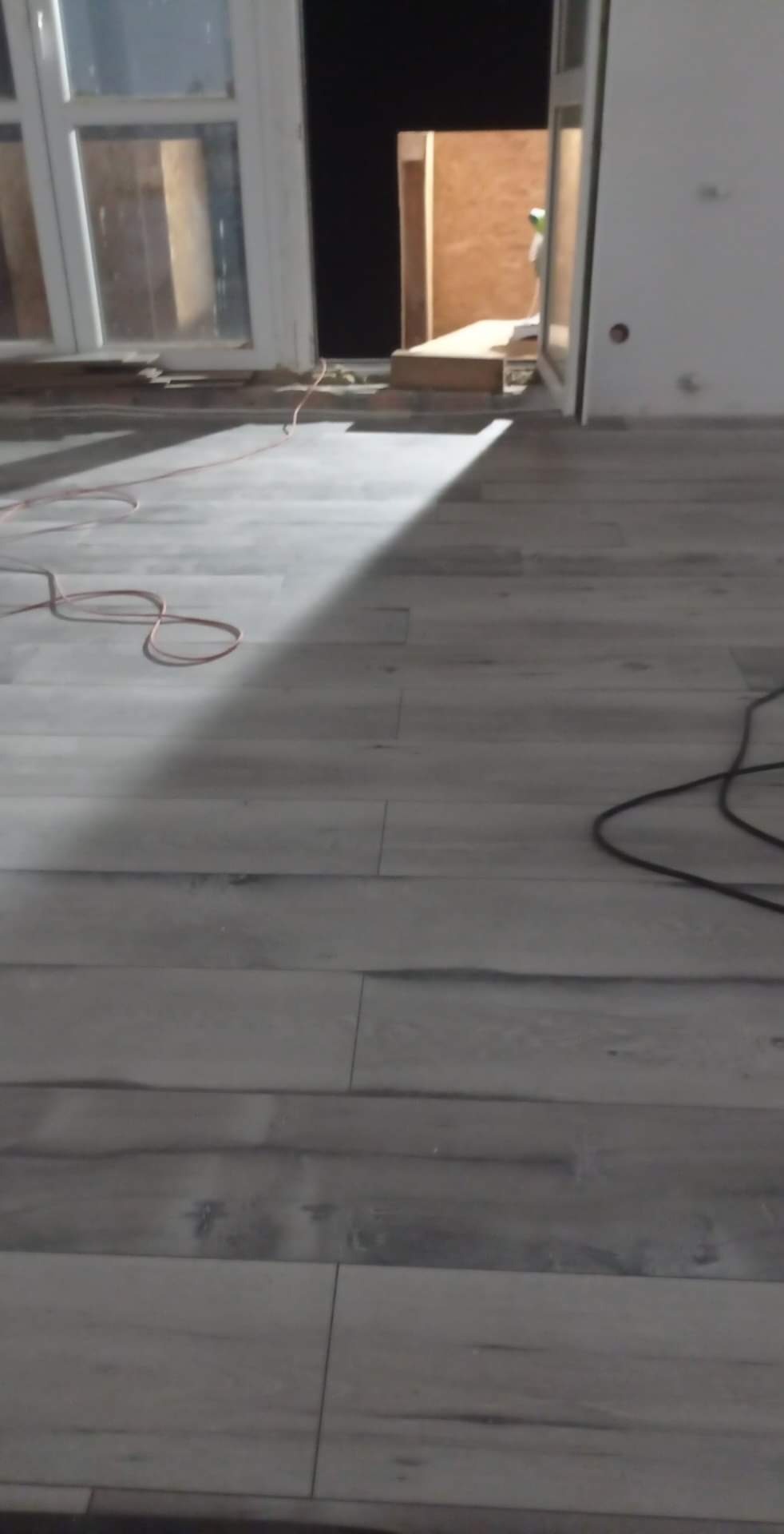 Ułożone panele podłogowe w odcieniach szarości, widoczne kable i fragment okna w trakcie prac wykończeniowych.